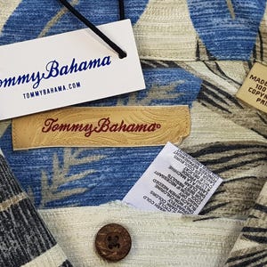 May include: A beige and blue patterned shirt with a Tommy Bahama label. The shirt has a button-down collar and short sleeves. The label says "Tommy Bahama" and "TOMMYBAHAMA.COM". The shirt also has a care label with instructions for washing and drying.