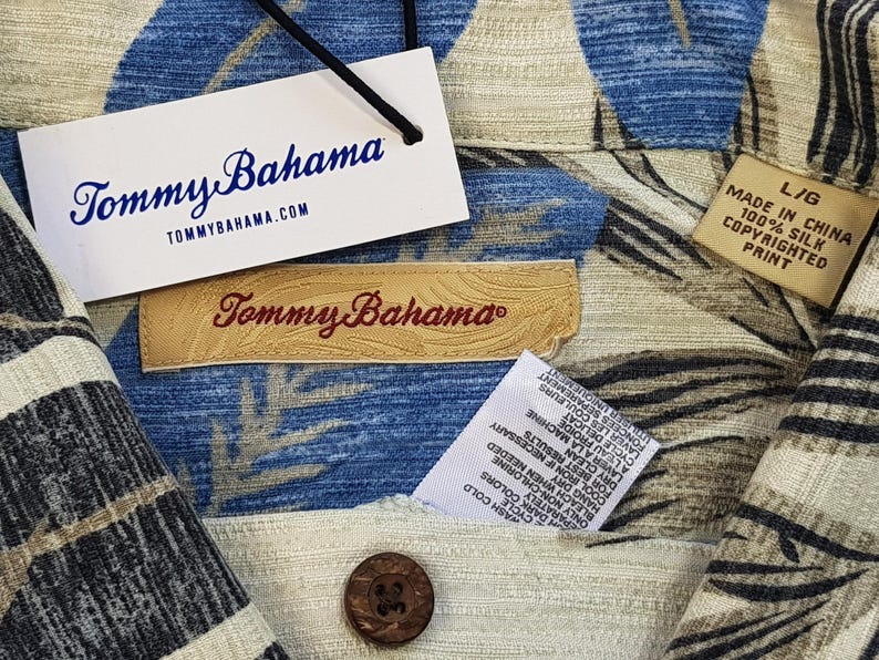 May include: A beige and blue patterned shirt with a Tommy Bahama label. The shirt has a button-down collar and short sleeves. The label says "Tommy Bahama" and "TOMMYBAHAMA.COM". The shirt also has a care label with instructions for washing and drying.