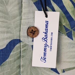 Puede incluir: Una etiqueta blanca con texto azul que dice "Tommy Bahama" y "tommybahama.com". La etiqueta est&aacute; unida a una camisa con estampado verde y azul con un bot&oacute;n de coco.