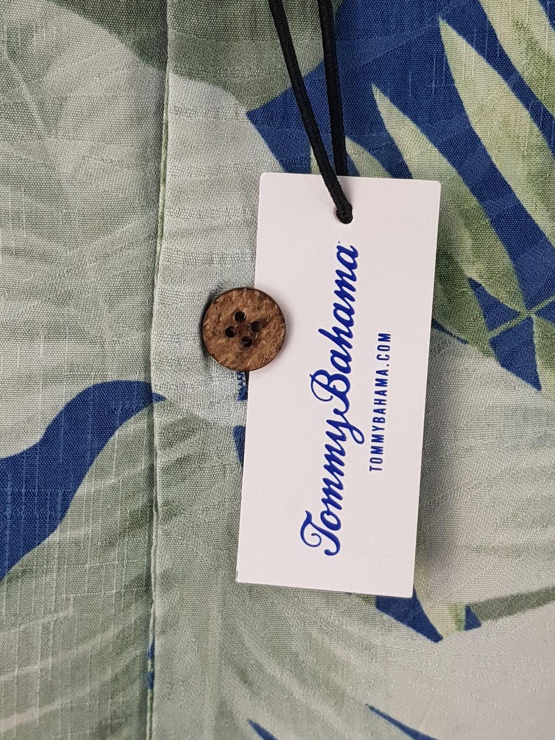 Puede incluir: Una etiqueta blanca con texto azul que dice "Tommy Bahama" y "tommybahama.com". La etiqueta est&aacute; unida a una camisa con estampado verde y azul con un bot&oacute;n de coco.