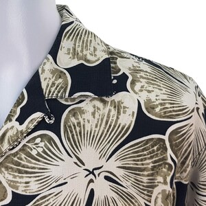 Peut inclure: Gros plan d'une chemise noire boutonn&eacute;e avec un motif floral. La chemise pr&eacute;sente de grandes fleurs stylis&eacute;es dans des tons beige et vert olive sur un fond sombre. Le col et le d&eacute;tail des boutons sont visibles.