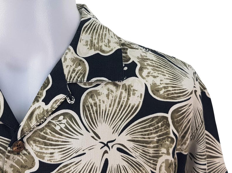 Peut inclure: Gros plan d'une chemise noire boutonn&eacute;e avec un motif floral. La chemise pr&eacute;sente de grandes fleurs stylis&eacute;es dans des tons beige et vert olive sur un fond sombre. Le col et le d&eacute;tail des boutons sont visibles.