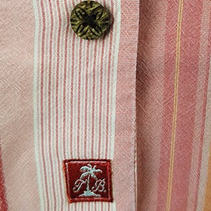 May include: A pink and white striped button-down shirt with a brown wooden button and a red embroidered patch with a palm tree and the letters "T&B".