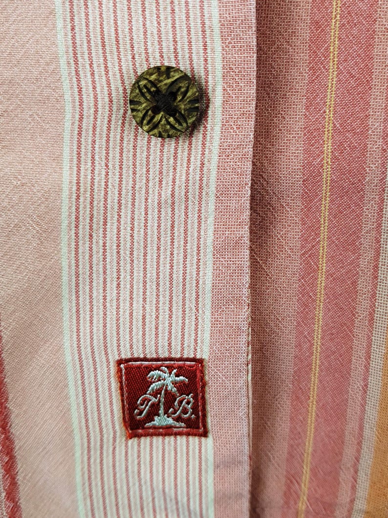 May include: A pink and white striped button-down shirt with a brown wooden button and a red embroidered patch with a palm tree and the letters "T&B".