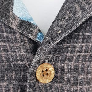 K&ouml;nnte beinhalten: Nahaufnahme eines schwarzen und grauen gemusterten Hemdes mit einem Holzknopf. Der Knopf hat ein kreuzf&ouml;rmiges Design.