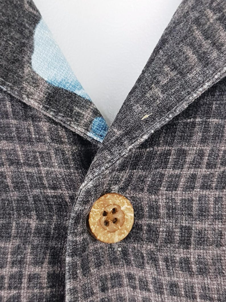 K&ouml;nnte beinhalten: Nahaufnahme eines schwarzen und grauen gemusterten Hemdes mit einem Holzknopf. Der Knopf hat ein kreuzf&ouml;rmiges Design.