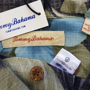K&ouml;nnte beinhalten: Nahaufnahme eines Tommy Bahama Hemdes mit einem floralen Muster in Blau-, Gr&uuml;n- und Schwarzt&ouml;nen. Das Hemd hat ein Etikett mit der Aufschrift "Tommy Bahama" und "Hergestellt in China 95% Seide 5% Andere Fasern".