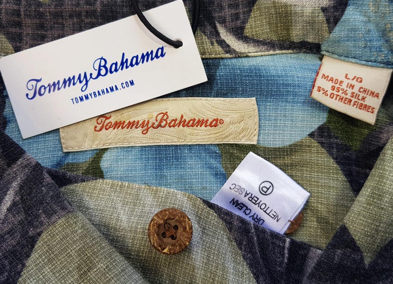 K&ouml;nnte beinhalten: Nahaufnahme eines Tommy Bahama Hemdes mit einem floralen Muster in Blau-, Gr&uuml;n- und Schwarzt&ouml;nen. Das Hemd hat ein Etikett mit der Aufschrift "Tommy Bahama" und "Hergestellt in China 95% Seide 5% Andere Fasern".