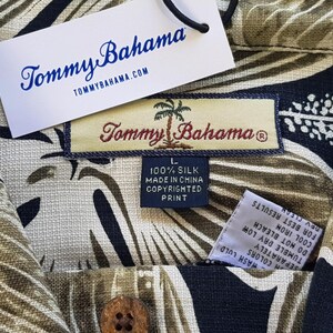 Peut inclure: Gros plan sur une chemise en soie Tommy Bahama avec un motif de feuilles tropicales beige, noir et vert. La chemise a une fermeture &agrave; boutons et une &eacute;tiquette indiquant "100% Soie Fabriqu&eacute; en Chine Copyrighted Print."