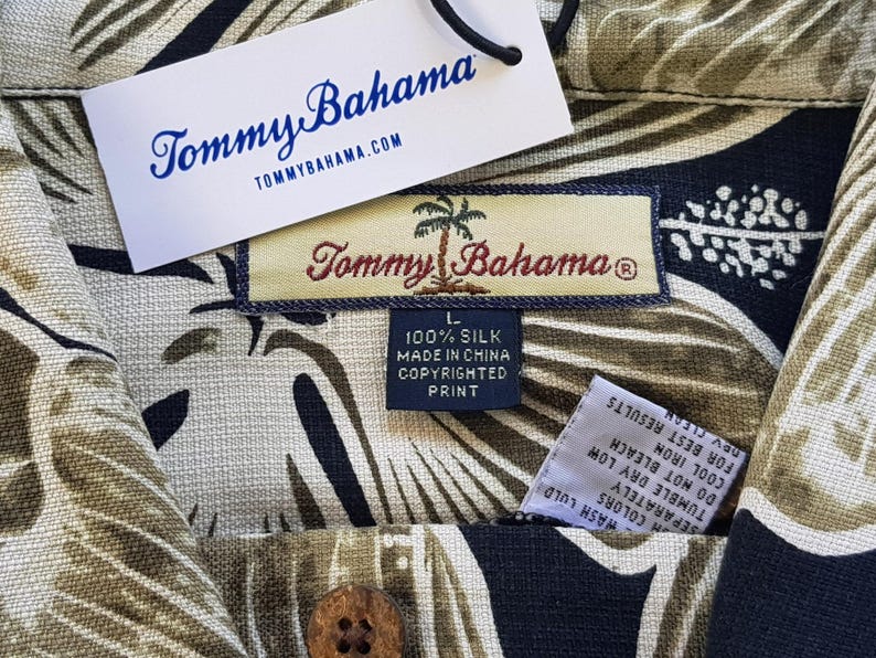 Peut inclure: Gros plan sur une chemise en soie Tommy Bahama avec un motif de feuilles tropicales beige, noir et vert. La chemise a une fermeture &agrave; boutons et une &eacute;tiquette indiquant "100% Soie Fabriqu&eacute; en Chine Copyrighted Print."