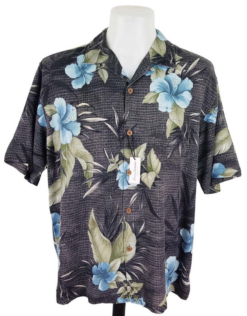 K&ouml;nnte beinhalten: Ein schwarzes, kurz&auml;rmeliges Hemd mit Kn&ouml;pfen und einem tropischen Muster mit blauen Hibiskusbl&uuml;ten und gr&uuml;nen Bl&auml;ttern. Das Hemd hat eine gewebte Textur und einen Kragen.