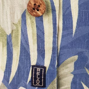 Puede incluir: Primer plano de una camisa de patr&oacute;n azul claro y verde con un bot&oacute;n de madera y una etiqueta negra que dice "RELAX".