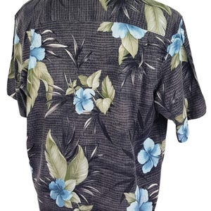 K&ouml;nnte beinhalten: Ein kurz&auml;rmeliges Hemd mit Kn&ouml;pfen und tropischem Muster. Das Hemd ist schwarz mit blauen und gr&uuml;nen Blumen und Bl&auml;ttern.