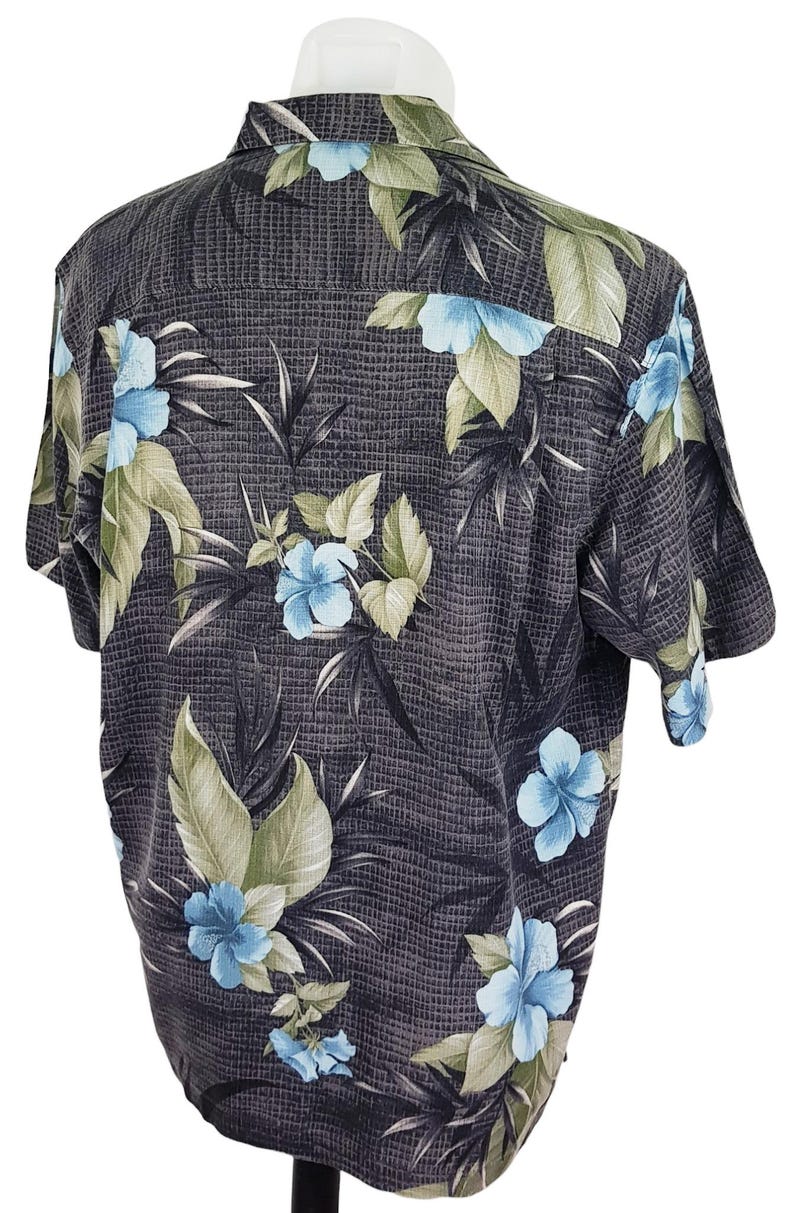 K&ouml;nnte beinhalten: Ein kurz&auml;rmeliges Hemd mit Kn&ouml;pfen und tropischem Muster. Das Hemd ist schwarz mit blauen und gr&uuml;nen Blumen und Bl&auml;ttern.