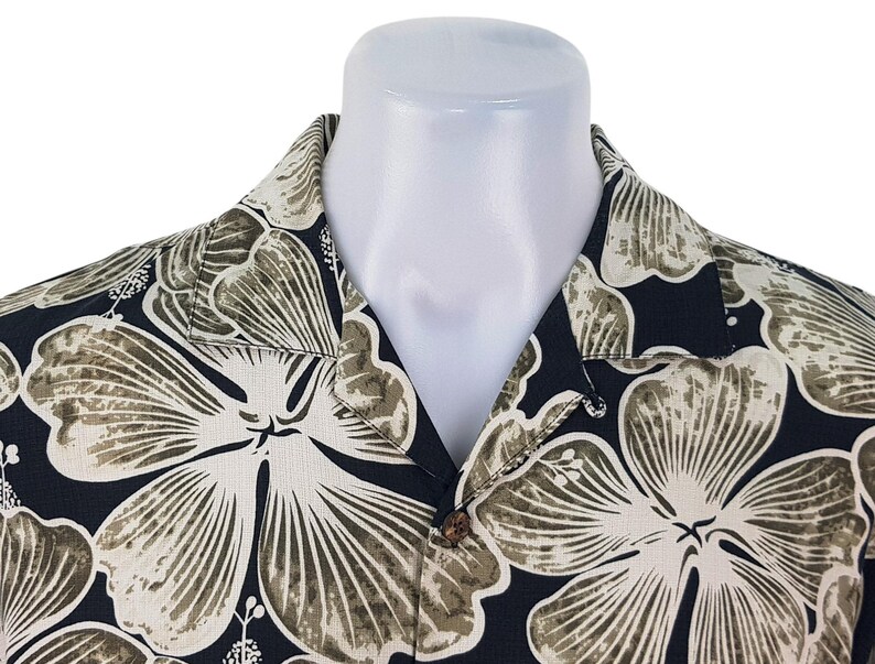 Peut inclure: Une chemise noire &agrave; boutons avec un grand motif floral dans les tons cr&egrave;me et vert olive. La chemise est pr&eacute;sent&eacute;e sur un mannequin blanc, mettant en valeur le col et les boutons. Le motif floral est pro&eacute;minent.