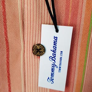 May include: A white tag with blue text that reads "Tommy Bahama" and "tommybahama.com" hanging from a black string. The tag is on a striped fabric background with red, orange, and white stripes.