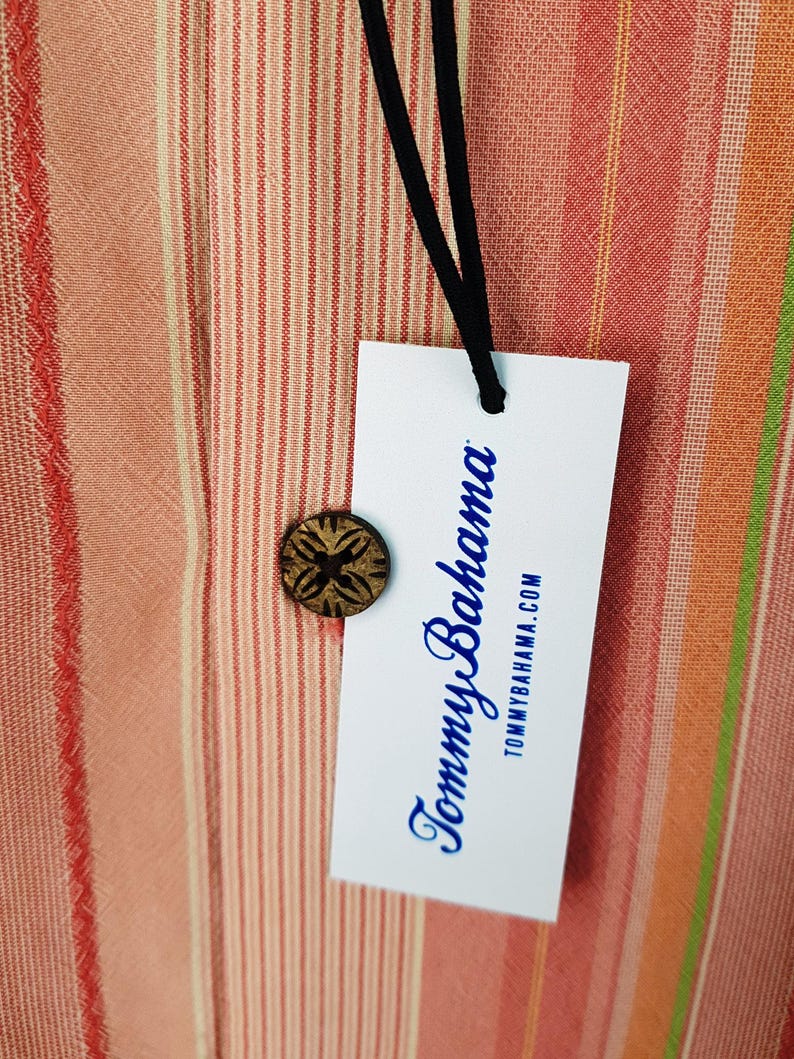 May include: A white tag with blue text that reads "Tommy Bahama" and "tommybahama.com" hanging from a black string. The tag is on a striped fabric background with red, orange, and white stripes.