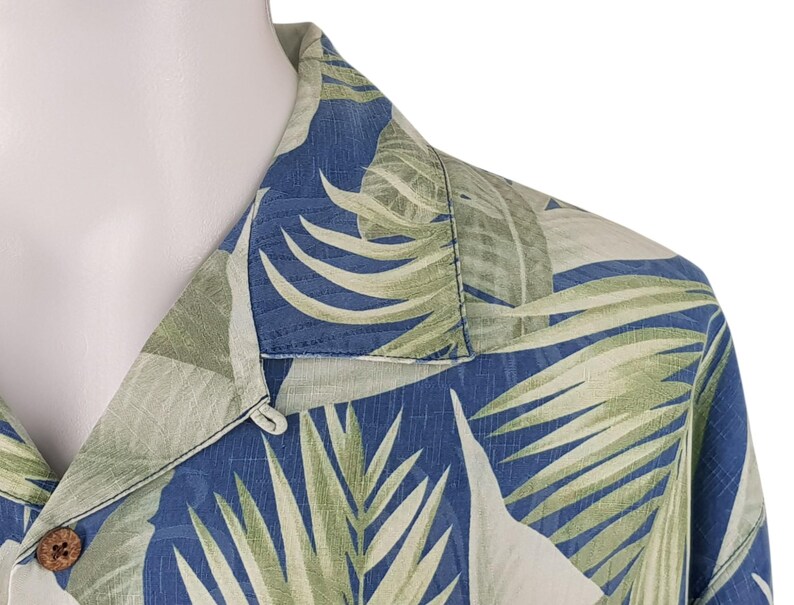 Puede incluir: Una camisa de botones azul y verde con cuello. La camisa tiene un dise&ntilde;o de hojas tropicales.
