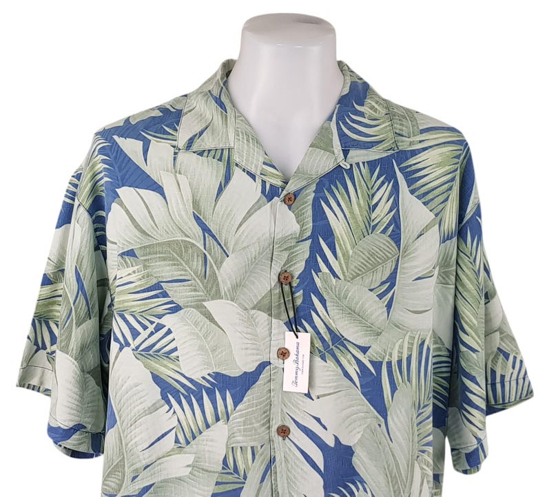 Puede incluir: Una camisa de manga corta con botones azul y verde con estampado de hojas tropicales. La camisa tiene cuello y cierre de botones. La camisa est&aacute; hecha de una tela ligera y es perfecta para el clima c&aacute;lido.