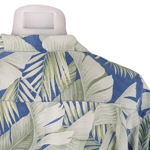 Puede incluir: Una camisa de estampado azul y verde con un dise&ntilde;o de hojas tropicales. La camisa est&aacute; hecha de una tela ligera y tiene un corte holgado.