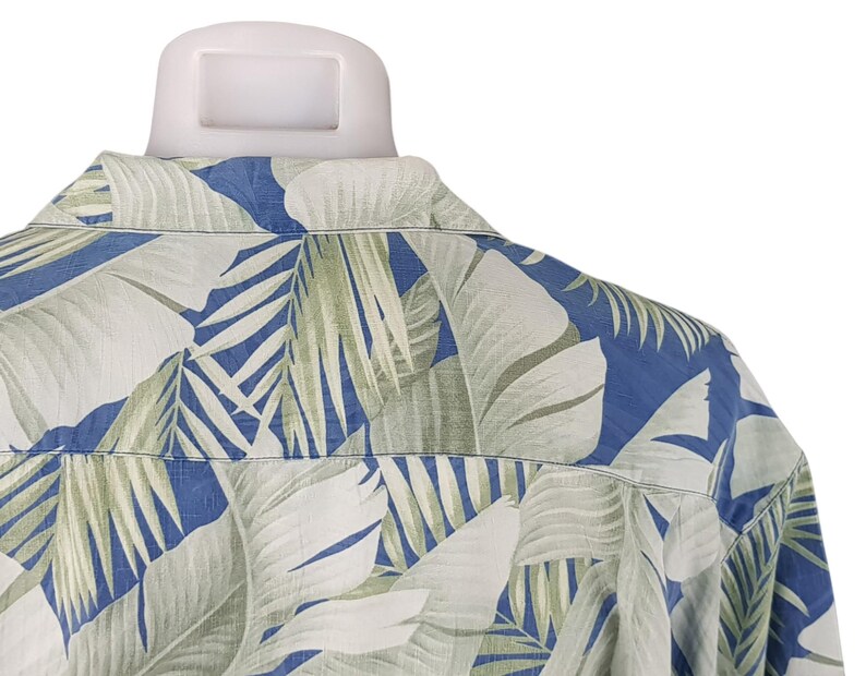 Puede incluir: Una camisa de estampado azul y verde con un dise&ntilde;o de hojas tropicales. La camisa est&aacute; hecha de una tela ligera y tiene un corte holgado.