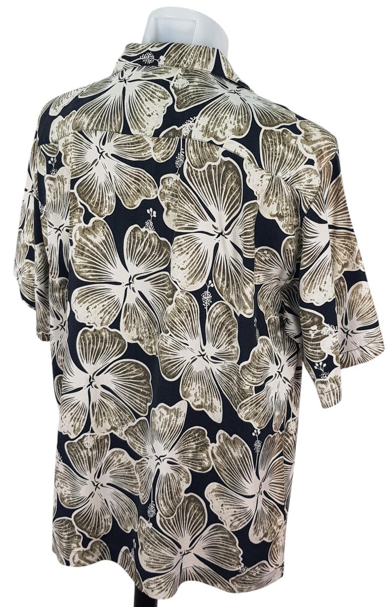 Peut inclure: Chemise &agrave; manches courtes avec un motif floral. La chemise pr&eacute;sente de grandes fleurs d'hibiscus stylis&eacute;es en blanc et marron sur un fond bleu marine fonc&eacute;. La chemise est pr&eacute;sent&eacute;e sur un mannequin.