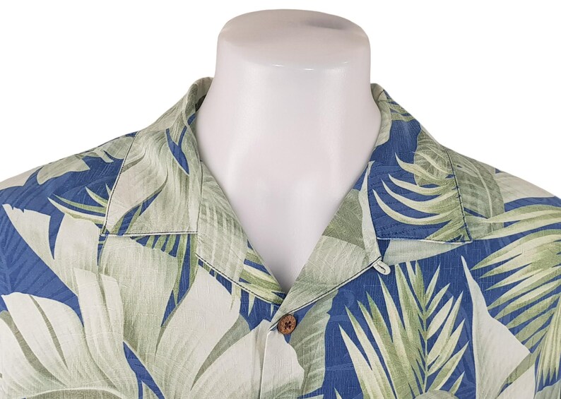 Puede incluir: Una camisa de estampado tropical azul y verde con cuello abotonado y un solo bot&oacute;n de cierre. La camisa tiene un corte holgado y est&aacute; hecha de una tela ligera.