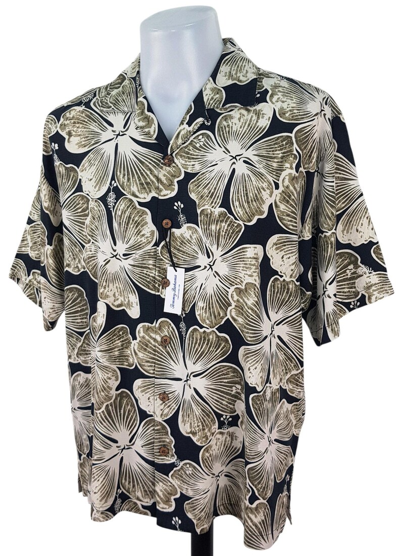 Peut inclure: Chemise &agrave; manches courtes avec un fond noir et un grand motif floral en blanc et vert olive. La chemise a un col classique et une &eacute;tiquette attach&eacute;e. Boutonn&eacute;e.