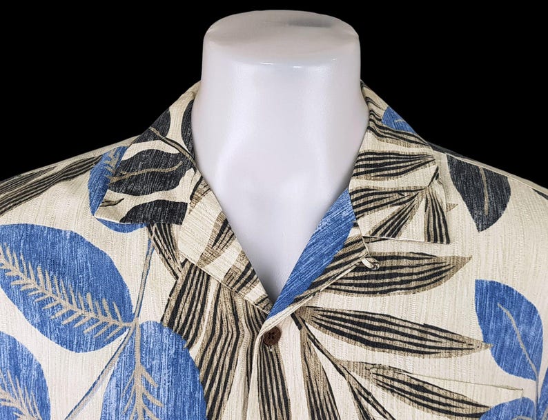 May include: A beige button-up shirt with a tropical print featuring blue, black, and brown leaves. The shirt has a collar and a button closure.