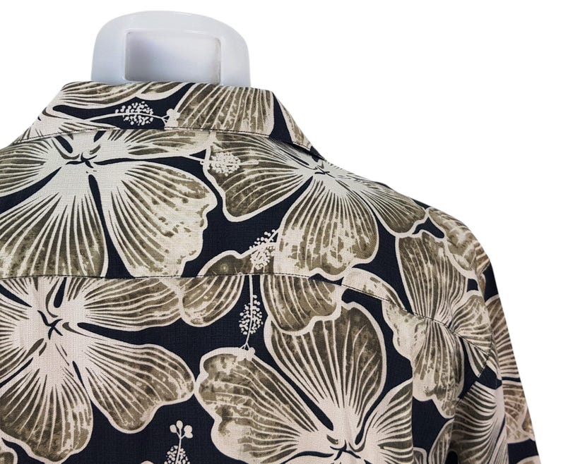 Peut inclure: Gros plan d'une chemise boutonn&eacute;e avec un grand motif floral. La chemise pr&eacute;sente de grandes fleurs stylis&eacute;es dans des tons beige et marron sur un fond bleu marine fonc&eacute;. Le col et le dos de la chemise sont visibles.