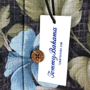 K&ouml;nnte beinhalten: Ein wei&szlig;es Etikett mit dem Text "Tommy Bahama" in blauer Schrift und der Website-Adresse "tommybahama.com" darunter. Das Etikett ist an einem schwarzen und gr&uuml;nen floralen Stoff mit einem Kokosnuss-Knopf befestigt.