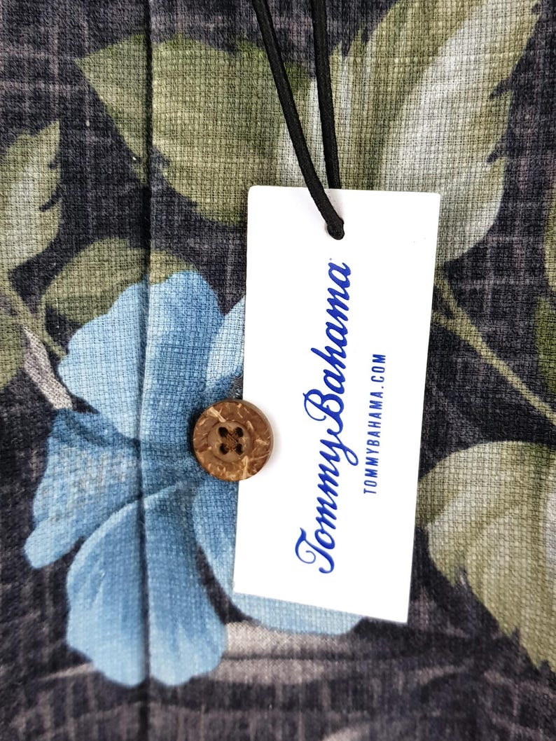 K&ouml;nnte beinhalten: Ein wei&szlig;es Etikett mit dem Text "Tommy Bahama" in blauer Schrift und der Website-Adresse "tommybahama.com" darunter. Das Etikett ist an einem schwarzen und gr&uuml;nen floralen Stoff mit einem Kokosnuss-Knopf befestigt.