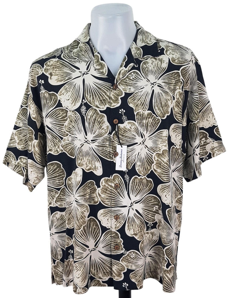 Peut inclure: Chemise &agrave; manches courtes boutonn&eacute;e avec un fond bleu marine fonc&eacute; et un grand motif floral dans les tons beige et vert olive. La chemise a un col classique et des boutons en bois. Une &eacute;tiquette avec le nom de la marque est attach&eacute;e.