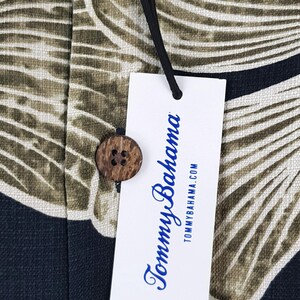 Peut inclure: Gros plan sur une &eacute;tiquette de chemise Tommy Bahama et un bouton en bois. L'&eacute;tiquette blanche porte le nom de la marque en &eacute;criture bleue et l'adresse du site web. Le tissu de la chemise pr&eacute;sente un motif de feuilles tropicales vertes, beiges et bleu marine.