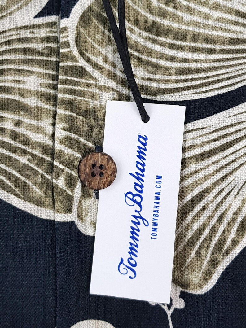 Peut inclure: Gros plan sur une &eacute;tiquette de chemise Tommy Bahama et un bouton en bois. L'&eacute;tiquette blanche porte le nom de la marque en &eacute;criture bleue et l'adresse du site web. Le tissu de la chemise pr&eacute;sente un motif de feuilles tropicales vertes, beiges et bleu marine.