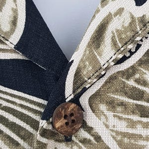 Peut inclure: Gros plan sur un col de chemise boutonn&eacute;e. Le tissu pr&eacute;sente un motif de feuilles tropicales dans des tons de noir, vert olive et cr&egrave;me. Un bouton rond marron est centr&eacute; sur le col. Le tissu de la chemise a un aspect textur&eacute;.