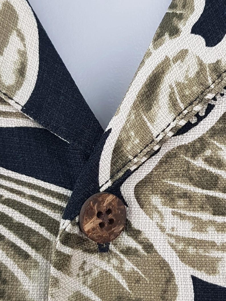Peut inclure: Gros plan sur un col de chemise boutonn&eacute;e. Le tissu pr&eacute;sente un motif de feuilles tropicales dans des tons de noir, vert olive et cr&egrave;me. Un bouton rond marron est centr&eacute; sur le col. Le tissu de la chemise a un aspect textur&eacute;.