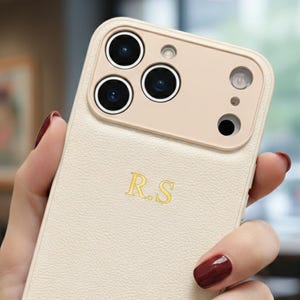 May include: A cream-colored phone case with a textured finish. The case has a beige camera module and gold-colored initials "R.S." The case is designed for a smartphone and features multiple camera cutouts.