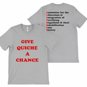 Op de afbeelding: Een lichtgrijs t-shirt met de tekst "GIVE QUICHE A CHANCE" in rode letters. De achterkant van het shirt toont het acroniem "CLITORIS" met de volledige zin "Committee for the Liberation & Integration of Terrifying Organisms & their Rehabilitation Into Society."