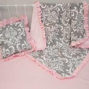 May include: A gray and white damask patterned baby blanket with pink ruffled trim. The blanket is folded and placed on a pink crib sheet with a matching gray and white damask patterned pillow with pink ruffled trim.