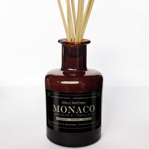 Könnte beinhalten: Eine dunkelbraune Glasflasche mit einem schwarzen Etikett, auf dem "Schön & Reich Edition MONACO 43°43'52.11" N - 7°25'11.13" E BERGAMOTTE - ORCHIDEE - VELICHES HANDGEFERTIGT IN DEUTSCHLAND SO-RICHT-GIF-WELL steht. Die Flasche enthält einen Satz natürlicher Holzstäbchen.