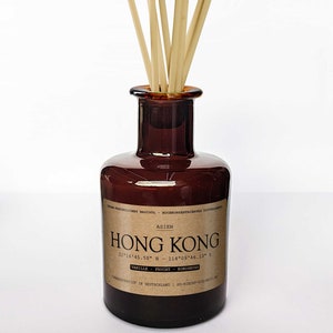 Könnte beinhalten: Eine braune Glasflasche mit einem braunen Etikett, auf dem "ASIEN HONG KONG 22°16'45.58" N - 114°09'46.13" E VANILLE FRUCHT KOKO" und "HERGESTELLT IN DEUTSCHLAND / SO-RICHT-DIE-WELT" steht. Die Flasche enthält einen Satz brauner Holzstäbchen.