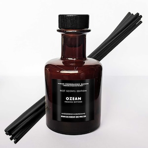 Könnte beinhalten: Eine braune Glasflasche mit einem schwarzen Etikett, auf dem "BEST SCENTS EDITION OZEAN PREMIUM DIFFUSER HANDGEFERTIGT IN DEUTSCHLAND WWW.SO-RICHT-DIE-WELT.DE" steht. Die Flasche ist mit einer klaren Flüssigkeit gefüllt und hat schwarze Stäbchen, die aus der Oberseite herausragen.