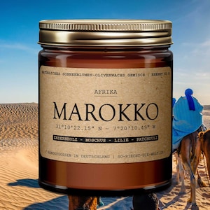 Puede incluir: Un tarro de vela de vidrio marrón con una tapa dorada. La etiqueta es marrón con texto blanco que dice "NATÜRLICHES SONNENBLUMEN-OLIVENWACHS GEMISCH | BRENNT 60 h", "AFRIKA", "MAROKKO", "31°10'22.15" N - 7°20'10.49" W", "ZEDERNHOLZ - MOSCHUS - LILIE - PATCHOULI" y "HANDGEGOSSEN IN DEUTSCHLAND | SO-RIECHT-DIE-WELT.DE".
