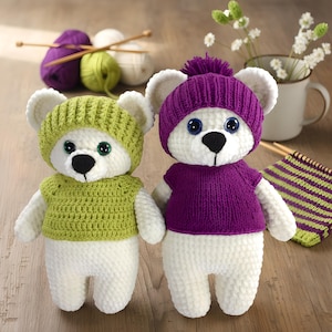 May include: Two handmade white teddy bears, each wearing a knitted hat and sweater. One bear has a green hat and sweater, the other a purple hat and sweater. Both have black noses and blue eyes. Knitting supplies are in the background.
