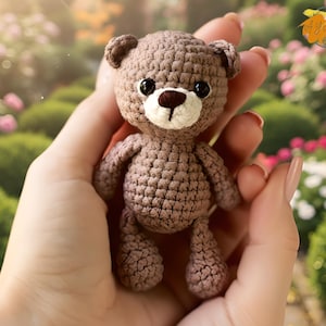 May include: A small, handmade crochet teddy bear in shades of brown, held in a person's hand. The bear has black eyes, a white muzzle, and a brown nose. The background is a blurred view of a garden with pink and green foliage.