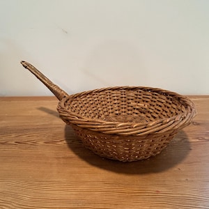May include: A woven basket with a long handle. The basket is made of light brown wicker and has a rounded shape. The handle extends from the side of the basket, creating a scoop-like design. The basket sits on a wooden surface.