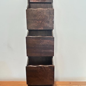May include: A dark brown wooden wall-mounted organizer with three compartments and a small key rack. Each compartment features a decorative swirl design. The organizer is made of wood and is designed to hold mail or other small items.
