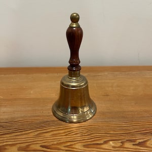 May include: A brass bell with a wooden handle. The bell is gold in color and has a rounded shape. The handle is dark brown and has a rounded knob on top.