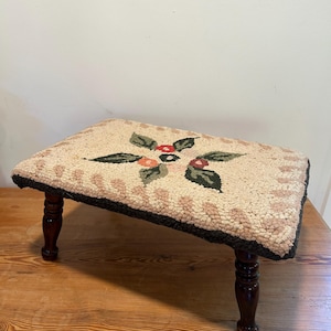 May include: A small, rectangular footstool with a beige, floral-patterned top and dark brown, turned wooden legs. The top features a central floral design with green leaves and red and orange berries, and a black border.